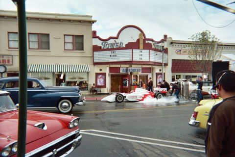 1990 Ford Bronco - Toyota-branded open-wheel Indy-style race car sits on a wet downtown street outside the Tower theater, flanked by 1950s-era cars and onlook…