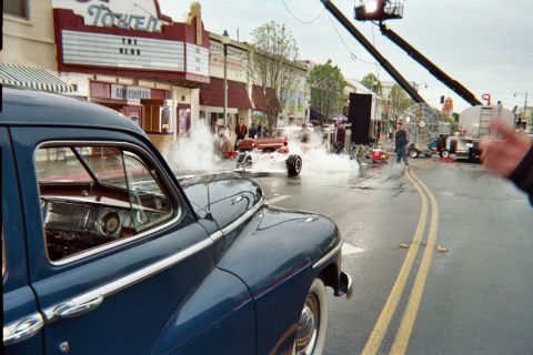 1990 Ford Bronco - dark blue vintage sedan dominates the foreground while a red-and-white race car emits smoke and a camera crane reaches over a wet street.
