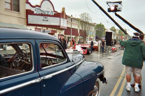 1990 Ford Bronco - polished dark blue 1950s sedan is parked along a wet street as a film crew operates a camera crane near an open-wheel race car by the Tower…