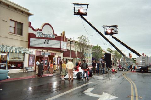 1990 Ford Bronco - Two camera cranes with bright lights tower over a wet main street where crew and period-dressed extras surround an open-wheel race car at t…
