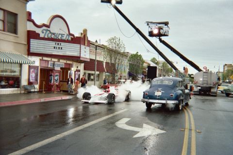 1990 Ford Bronco - red-and-white open-wheel race car spins up tire smoke next to a dark vintage sedan on a wet street with a camera crane overhead at the Towe…