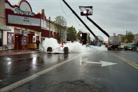 1990 Ford Bronco - red-and-white open-wheel race car sits enveloped in tire smoke on a wet downtown street with a camera crane and fan staged nearby.
