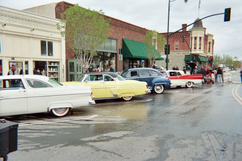 1990 Ford Bronco - lineup of 1950s automobiles in white, yellow, blue and red is parked along a wet downtown street fronted by old brick storefronts.