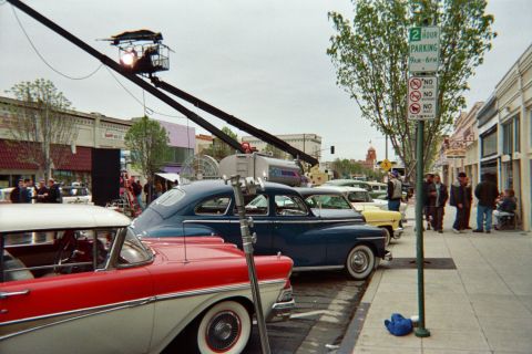 1990 Ford Bronco - Red, blue and yellow vintage cars line a curb under a camera crane, beside a green two-hour parking sign and gathered onlookers.