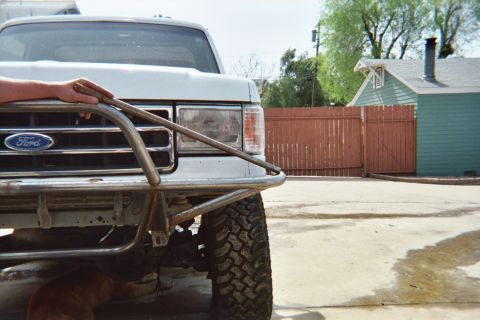 1990 Ford Bronco - close-up of a white Ford pickup's tube front bumper and grille, parked on concrete beside a red wooden fence and a green house.