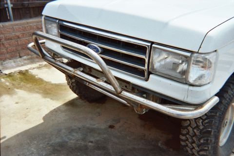 1990 Ford Bronco - close-up of a white Ford pickup's chrome grille, square headlight and tubular front bumper above a knobby off-road tire.