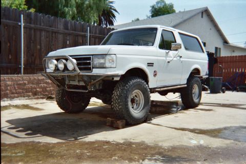 1990 Ford Bronco - white lifted Ford Bronco on off-road tires, fitted with a tube front bumper and round driving lights, sits in a fenced backyard.
