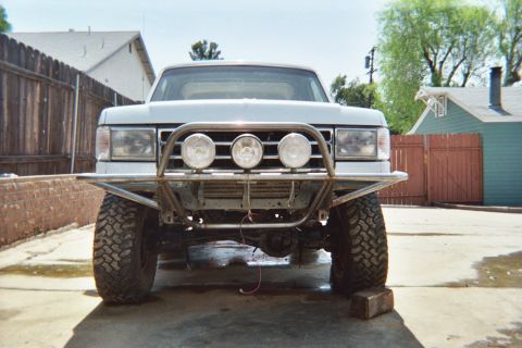 1990 Ford Bronco - head-on view of a white lifted Ford on big tires showing a tube front bumper with three round driving lights, parked on concrete.