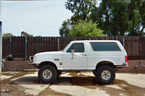 1990 Ford Bronco - side profile of a white lifted two-door Ford Bronco on big off-road tires, parked in a backyard beside a wooden fence.