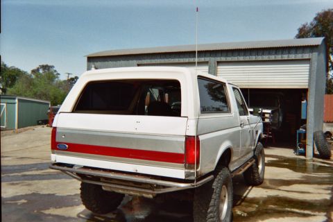 1990 Ford Bronco - rear three-quarter view of a white-and-red lifted Ford Bronco with a tall antenna, parked on concrete in front of an open metal shop.