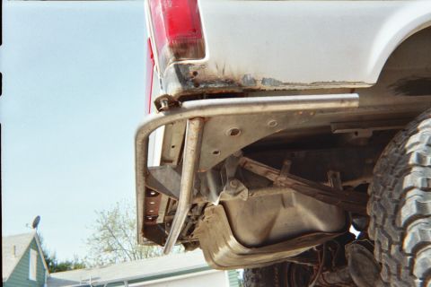 1990 Ford Bronco - close-up of the rear tube bumper, fuel tank and undercarriage beneath a white lifted truck's red taillight area.