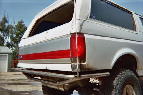 1990 Ford Bronco - rear corner of a white-and-red lifted Ford Bronco showing the taillight, side stripe and tube bumper, parked on concrete by a shed.