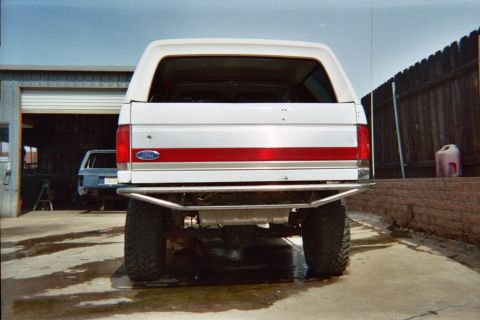 1990 Ford Bronco - straight-on rear view of a white-and-red lifted Ford Bronco with a tube bumper, parked on concrete between a metal shop and a brick wall.