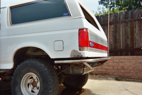 1990 Ford Bronco - rear quarter of a white lifted Ford Bronco showing the fuel door, red side stripe, taillight and tube bumper beside a wood fence.