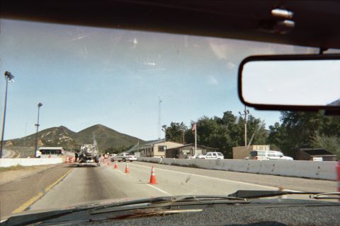 1990 Ford Bronco - Through-the-windshield shot of a highway inspection station with concrete barriers, traffic cones and a flag beside low buildings under haz…