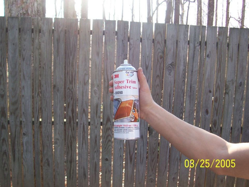 Hand holding a can of 3M Super Trim Adhesive 08090 in front of a wood fence
