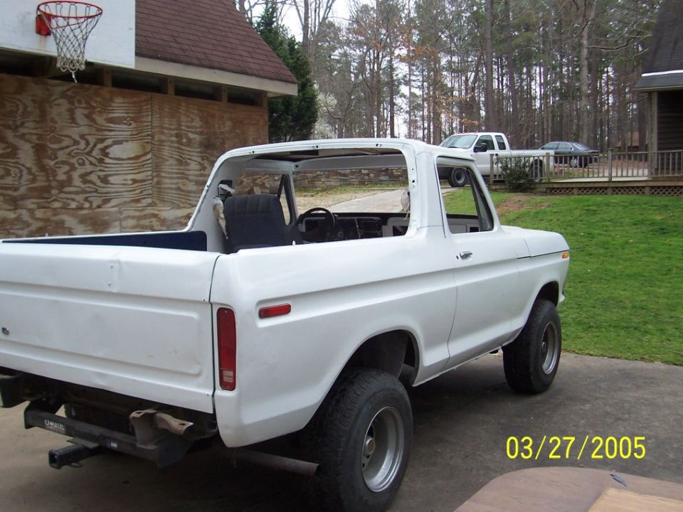 White Ford Bronco, top removed, parked outside a house. Another truck and car are visible in the background