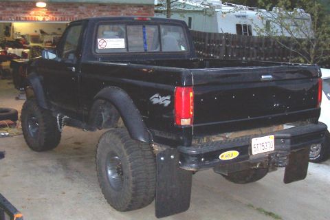 1986 Ford Bronco - This black pickup truck features oversized tires and mud flaps, seen from the rear.