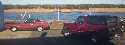 1971 Ford Mustang - Two red Ford vehicles, a Mustang convertible and a Bronco, are parked outdoors near a body of water.