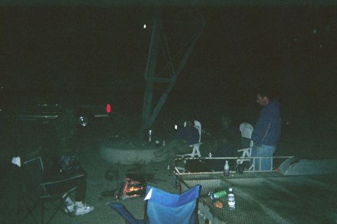 1990 Ford Bronco - dark nighttime scene of people seated in folding chairs around a small campfire near a tower structure, with drinks on a table.