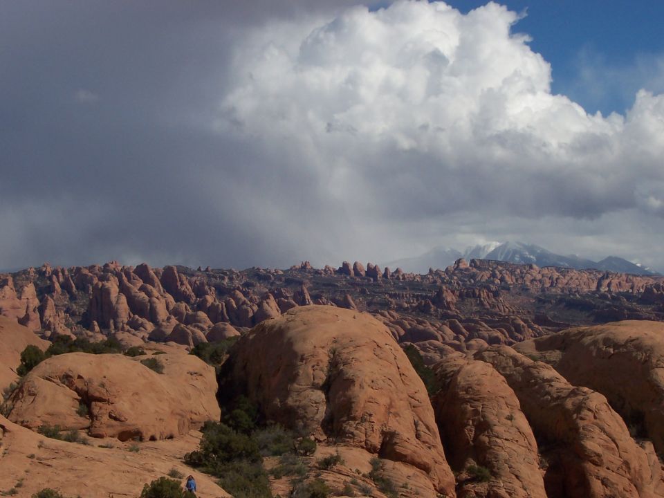 Desert landscape with large rock formations and dramatic storm clouds overhead. A person is visible on a rock in the lower left