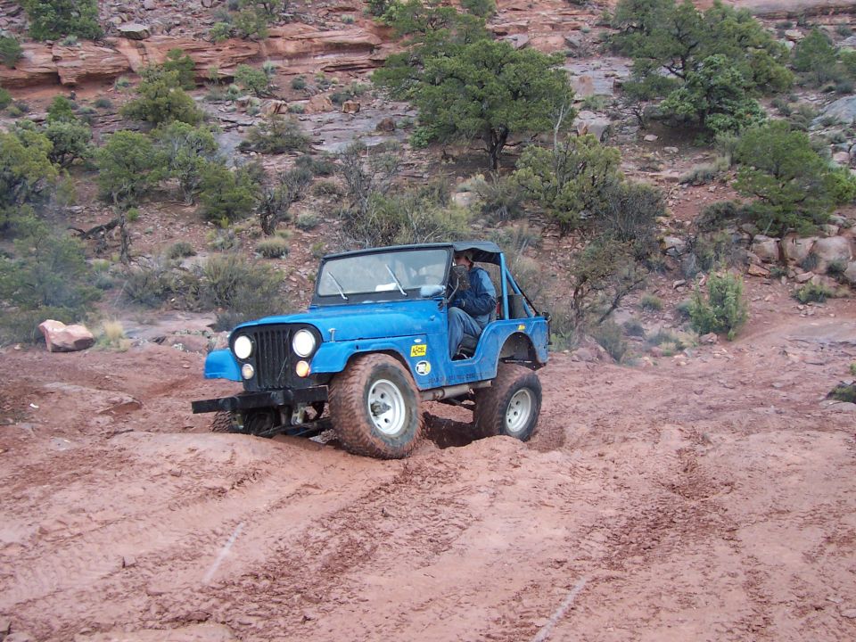 Blue Jeep CJ-7 navigating a muddy trail with red rock formations in background