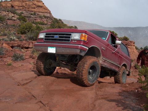 1991 Ford Bronco - A red Ford Bronco navigates a challenging off-road trail with steep inclines and rocky obstacles.