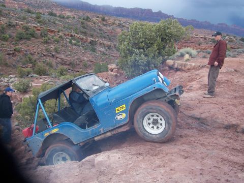 1991 Ford Bronco - A blue Jeep Wrangler navigates a difficult off-road obstacle in a desert landscape.