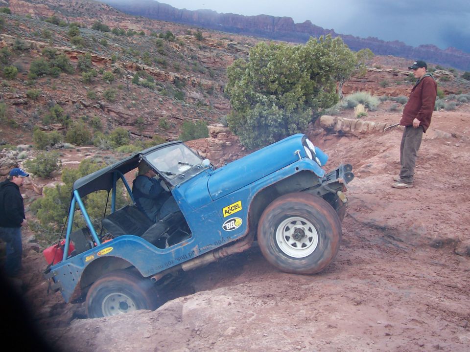 Blue Jeep Wrangler struggling on a rocky, muddy trail with two people watching