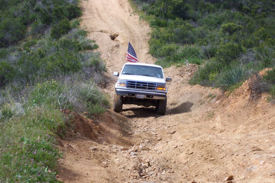 White Ford Bronco with American flag climbing a rutted dirt trail, front view