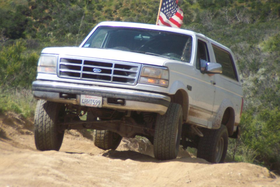 White Ford Bronco lifting a front wheel on a dirt trail, American flag mounted, plate 4HHT599