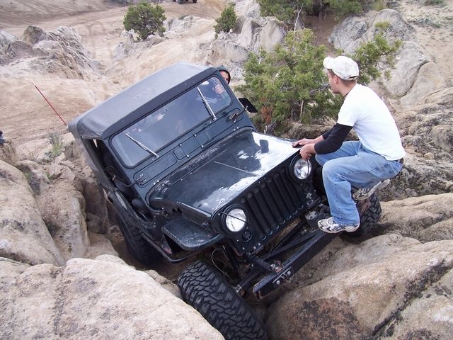 Black flat-fender Willys Jeep crawling down granite rock with a spotter alongside