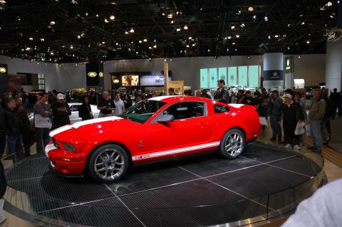 2005 Ford Mustang - A red Ford Mustang GT500 is displayed at an auto show with spectators around.