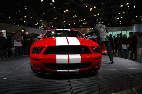 2005 Ford Mustang - A red 2005 Ford Mustang with white racing stripes is displayed on a platform, with a person interacting with the driver's side door.