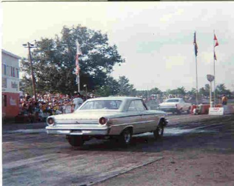 1963 Ford Galaxie - A light-colored car races down a drag strip with spectators in the background and flags overhead.