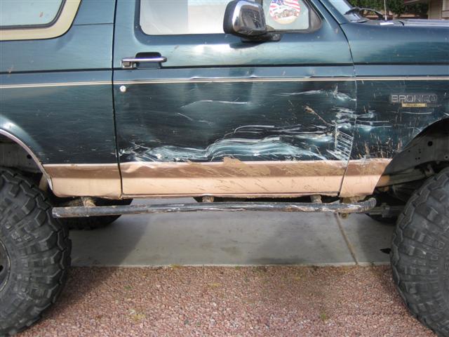Side view of a green Ford Bronco with scraped door and tan lower trim, parked on concrete