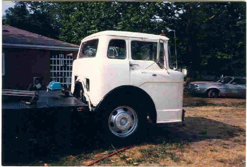 1957 Ford Deluxe - A white cab-over truck is positioned on a flatbed trailer, with another vehicle in the background.