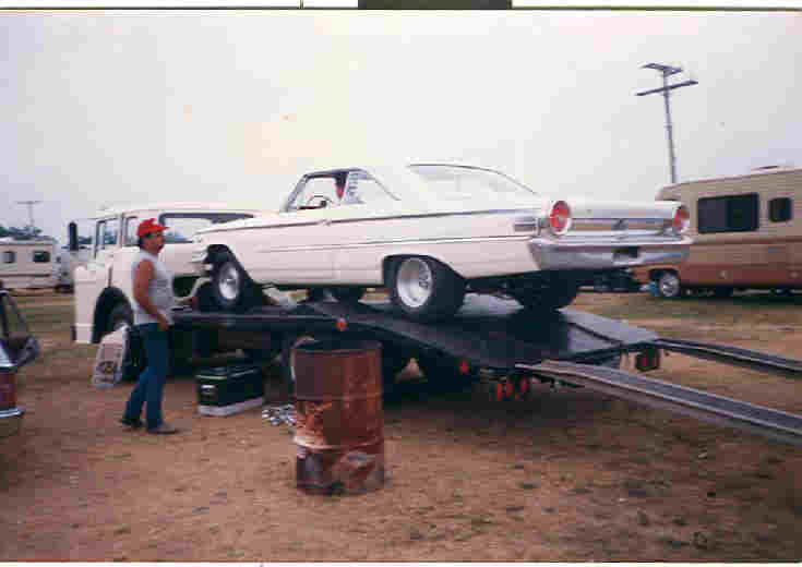White 1960s drag car loaded on a tilt-deck car hauler at a dirt racetrack pit