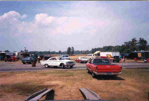 1957 Ford Deluxe - A drag strip scene with several cars and trailers under a partly cloudy sky.