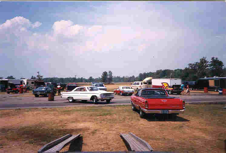 Red and white 1960s cars in drag strip pit area seen past car hauler ramps