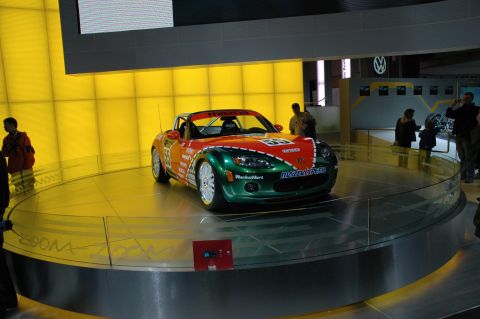 2005 Ford Mustang - Mazda Miata race car displayed at an auto show with a yellow background.