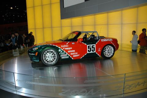 2005 Ford Mustang - A Mazda Miata race car is displayed on a rotating platform with a yellow illuminated background.