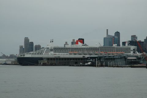 2005 Ford Mustang - The Queen Mary 2 cruise ship is docked in a harbor with a city skyline visible behind it.