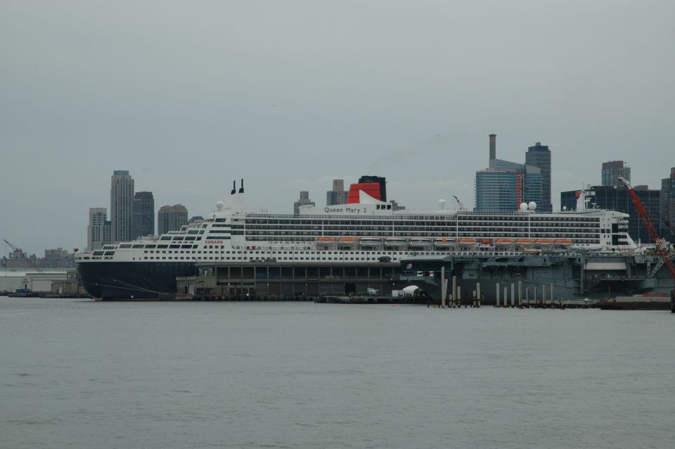 Queen Mary 2 cruise ship docked next to an aircraft carrier with city skyline in background