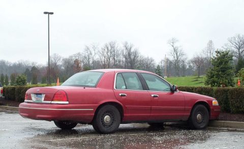 1998 Ford Crown Victoria - A red 1998 Ford Crown Victoria is parked on a wet surface with trees in the background.