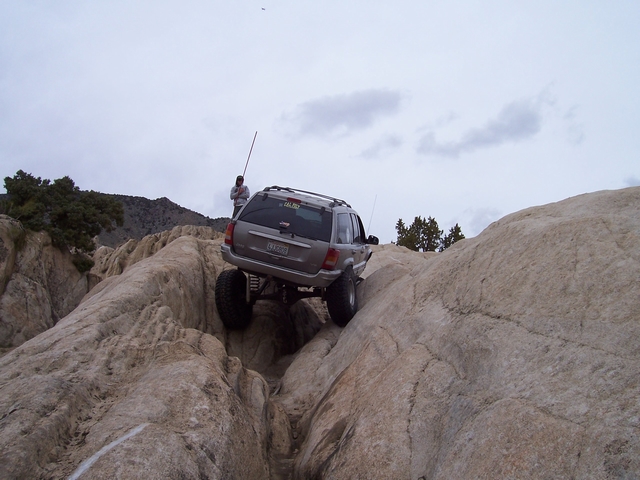 Jeep Grand Cherokee climbing over large rocks on an off-road trail. A person stands nearby holding a long pole