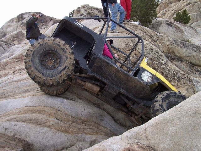 Off-road buggy with roll cage climbing steep rock formation. People watch from above