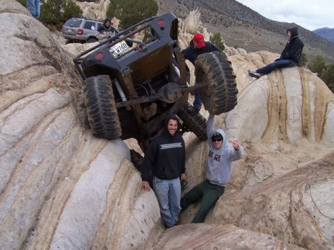 1989 Ford Bronco - An off-road vehicle is stuck on a rocky incline, with several people observing and assisting. Another SUV is visible in the background.