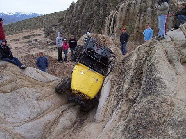 Yellow off-road vehicle tilted on its side in a rocky ravine, with people watching from above