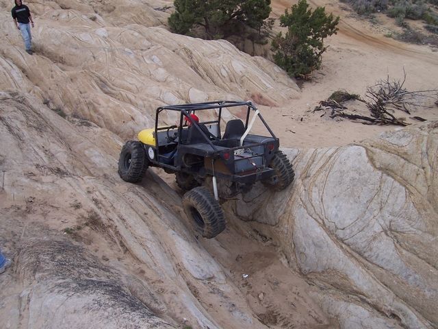 Yellow off-road buggy with a roll cage climbing a steep, sandy rock formation. Two people are visible, one standing on the rocks above and one partially visible at the bottom left